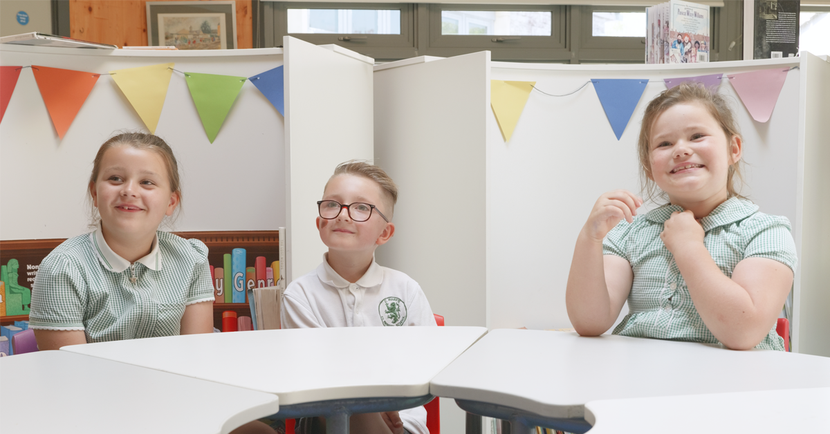 Three young children in school uniform sit around a table in a school library with bunting behind them