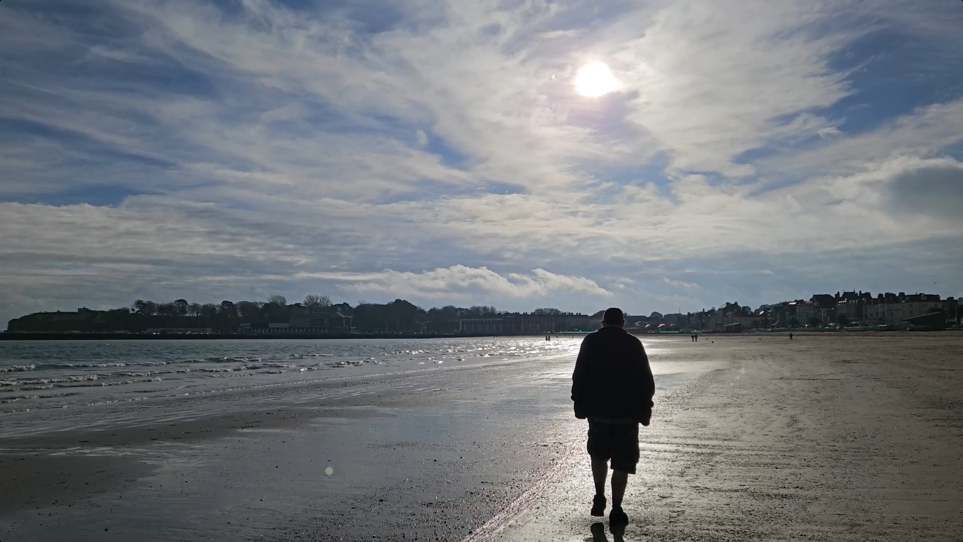 A man is walking away from the camera along a beach where the tide is out. The sea is to his left. Weymouth town centre is in front of him, in the distance.