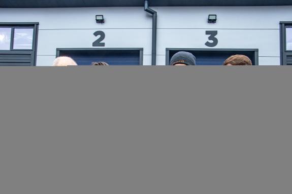 4 men stand outside of 2 business units, the units in the background have numbers 2 and 3 on them, they are grey, the 4 men in the foreground are wearing coats, one man is handing a key to another man which represents them taking on their new units